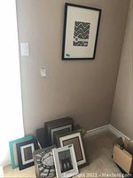Picture frames on floor corner, showing an assortment of sizes and finishes, mostly black and wood frames with glass fronts
