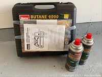 Carrying case labeled Home Butane 9000 with two butane fuel canisters and instruction manual taped on front.