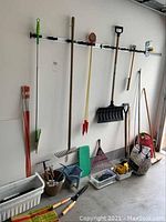 Photo of assorted garden tools hanging on wall and standing against wall including rakes, snow shovel, various hand tools in containers, planters and driveway markers.