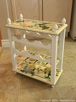 Front view of small wine rack showing painted finish and floral decoupage on top and bottom surfaces with four finials at corners.