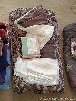 Folded queen comforter with brown and beige floral pattern, white folded linens set, and boxed new double bed linens on top.