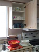 Nested set of melamine mixing bowls in red, orange, yellow, cream colors on counter; white Jamie Oliver serving bowls stacked; four Corelle 10.5 x 10.5 inch square plates on counter in front of stove.