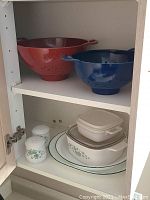 Four vintage CorningWare casserole dishes with lids stacked inside cupboard, two small ceramic spice jars on the shelf.