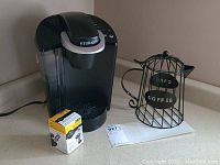 Front view of black and silver Keurig K40 coffee maker with reusable pod packaging and wire coffee pot decor on kitchen counter.