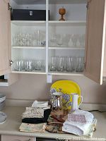 Open cupboard doors showing three shelves with various glassware items including old fashioned tumblers, highball glasses, wine glasses, and a wooden goblet on top shelf. Counter below the cupboard holds folded linens, two serving trays (yellow with floral design and one smaller with floral patterns), two ceramic pitchers (silver and white), and a small decorative glass bowl.