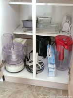 Photo of cabinet shelf containing Braun food processor with accessories, electric hand mixer, black electric kettle, Brita water pitcher with red lid, and box of filters.
