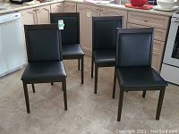 Photo shows four dining chairs with dark brown wood frames and black leather-look padded seats and backs arranged in kitchen.