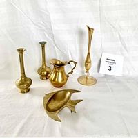 Five small brass items arranged showing two twin bud vases, mini pitcher, tall bud vase, and fish ashtray.