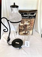 Photo of vintage glass wine fountain with black wrought iron stand and original box, displayed with auction card.