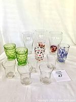 Front and top side view of 14 assorted vintage glasses including clear, green ribbed, and decorative printed glasses.