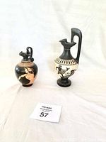 Photo showing two Greek-style vases on a white fabric background, front and side views visible.