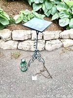 Full view of the black metal side table with a square glass top, green lantern, and basket hook arranged on ground outside in front of stone wall