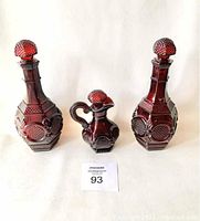 Front view of two tall octagonal cranberry glass decanters and one smaller pitcher with stoppers on top.