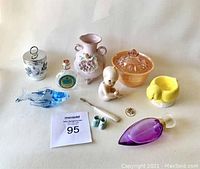 Photo showing assortment of vintage items including porcelain figurines, glass and ceramic pieces, bottles, and kitchen collectibles laid out on white surface.