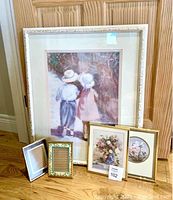 Overall view of five framed items leaning against a wooden surface: large Ivan Anderson print, two floral art prints, and two small photo frames.