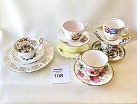Five assorted vintage teacup and saucer sets displayed together, showing varied floral and heraldic patterns with gold trim.