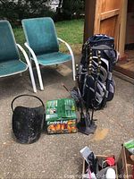Wide view of lot showing two green patio chairs, black metal fireplace tools with brass finials, CALINA golf bag, box of Enviro-Log fire starters, and black leather log carrier on pavement outside