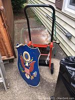 Metal wall plaque featuring the U.S. Eagle Great Seal on a blue shield-shaped background beside a vintage hand truck