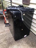 Full view of black GE mini fridge covered partially with black plastic wrapping, power cord draped over top, shown outdoors on concrete