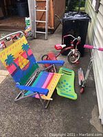 Photo showing the colorful foldable beach chair, green and blue Teeter Popper, red Radio flyer tricycle, and red and silver scooter all placed outdoors on concrete surface.