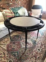 Full view of round poker table set up in a room, showing black leg supports and beige playing surface.