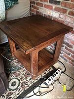 Solid wood side table photographed from an angle showing tabletop, legs, lower shelf, and portion of surrounding area.