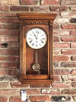 Front view of the wooden wall clock with visible pendulum and Roman numeral clock face against a brick wall.