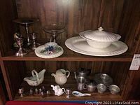 Photo of wooden cabinet shelves containing various silver-toned candlesticks, ceramic and metal serving ware including Johnson Brothers pieces and Italian tinware tea set.