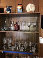 Shelf with assorted glassware and ceramics including a deep blue Royal Winton ceramic pitcher, a multicolor Japanese vase, and a white floral vase. Various stemware and candlesticks on lower shelves.
