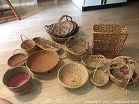 Photo showing a variety of woven baskets of different sizes and styles arranged on a floor, some with handles, some shallow, one with fabric cover.