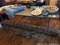 Full top and side view of the gray catering table with assorted household items on top and a patterned rug rolled underneath