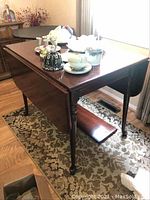 Cherry wood rectangular drop leaf table with items on top, one drop leaf folded down to the side, extra leaf shown leaning against wall, wooden legs visible.