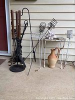 Photo showing two sets of fireplace tools with stands, white plant stands, spiral candle holders, clay pitcher and seashells on a concrete porch.