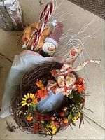 Photo showing artificial pine wreath with candy canes, a snowman figure in a knit hat, white decorative twigs, and a light brown pumpkin. Some items are in a plastic bag.
