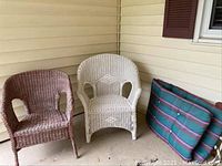 Two wicker chairs, one brown on the left and one white on the right, placed outside against a beige siding wall. Also includes a striped lounger pad with dark green, blue, and red stripes folded upright against the wall.