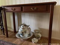 Front view of the cherry wood foyer table showing two drawers with brass handles and floral ceramic items on the floor beneath the table.