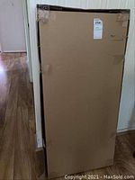Photo of the unopened IKEA Erik file cabinet in its tall rectangular cardboard box on a wooden floor.
