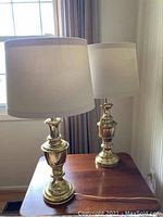 Pair of vintage brass-finished table lamps on wooden surface near window with white shades.