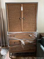 Front view of the entertainment center wrapped in plastic, showing wicker exterior with two doors on top and three drawers below.