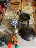 Kitchenware items arranged on a table including baked goods pans, Blue Willow serving dish, cast iron skillets, teapot, ceramic and wooden items, and vintage toys.