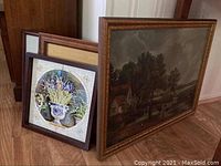 Photo showing four framed artworks leaning against a wall on a wooden floor, including a large painting and floral tile artworks.