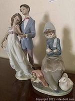 Two figurines side by side on wood surface; dancing couple at left, seated girl with geese at right
