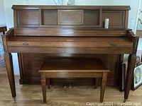 Full frontal view of Baldwin upright piano with matching bench underneath, showing wood finish, pedal hardware, and piano front grille.