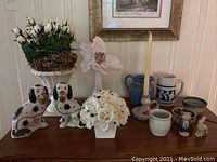 Overview of lot on wooden surface showing the two Staffordshire ceramic fireplace dogs, multiple faux flower arrangements, and various pieces of Williamsburg pottery including mugs and small bowls