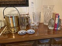 Photo showing brass ice bucket, two Delft ceramic plates, glass vases, glass elephant bookends, and books on wood cabinet.