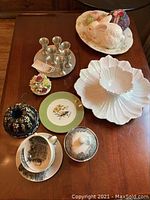 Wide view of all items on a polished wooden table showing hammered pewter plate with six liqueur glasses, Theodore Haviland floral platter, Coalport flower figurines, Maryland plate with bird design, ceramic Bundt pan with lid, two cups and saucers with canister marked Made in Italy.