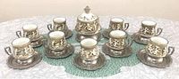 Photo showing all 9 espresso cups, 10 saucers, and sugar bowl arranged on table, highlighting the cherubs relief design and metal handles/bases.