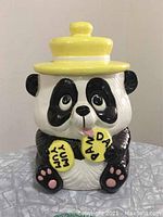 Front view of the ceramic panda cookie jar wearing a yellow hat and holding two yellow cookies.