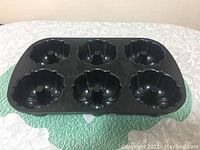 Black Nordic Ware muffin tray with six bundt-shaped molds on a green and white quilted surface.