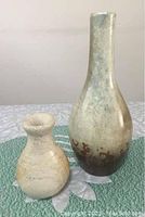 Tall ceramic pottery vase with narrow neck and brown base glaze, standing on cloth surface with green placemat, showing overall shape and wear